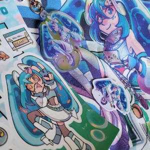 Könnte beinhalten: Ein Acryl-Schlüsselanhänger und zwei Aufkleber mit einer Anime-Figur mit türkisfarbenen Haaren, Schutzbrille und futuristischem Outfit. Der Schlüsselanhänger hat einen regenbogenfarbenen Metallverschluss. Ein Aufkleber enthält den Text Miku Miku... BEAM!!!!!.
