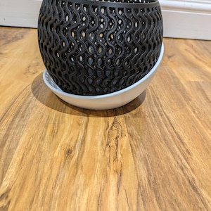 May include: A black, open-weave planter pot with a white saucer. The pot features a repeating, wavy design and rests on a white, round saucer. The pot and saucer are on a wood-grain surface.