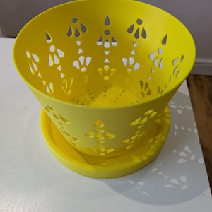 Puede incluir: Un cuenco decorativo amarillo brillante con un platillo a juego. El cuenco tiene una forma redondeada con intrincados patrones recortados en los lados. El cuenco y el platillo están hechos de un material sólido, probablemente plástico o cerámica.