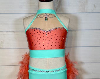 May include: A coral and turquoise dance costume with a matching headpiece. The halter top is coral with rhinestone embellishments and a turquoise neckline. The costume includes turquoise shorts and coral feather accents.