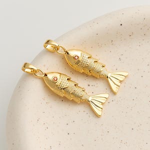 Peut inclure: Deux pendentifs dorés en forme de poisson, avec des écailles texturées et des détails d'yeux rouges. Chaque pendentif a une boucle en haut pour l'attacher à une chaîne. Les pendentifs sont sur une surface claire et mouchetée. Bijoux.