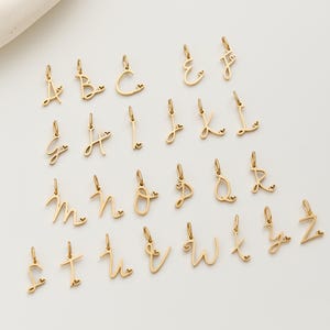 Könnte beinhalten: Eine Sammlung goldfarbener Buchstabenanhänger, jeweils mit einem kleinen Herzdetail. Die Buchstaben von A bis Z sind in Schreibschrift gehalten und auf einer weißen Oberfläche angeordnet. Ideal für personalisierten Schmuck.