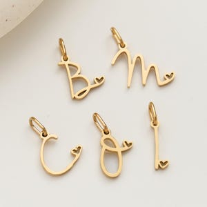 May include: Five gold-colored initial charms, each with a small heart detail. The letters are B, M, C, O, and I. Each charm has a small loop at the top for attaching to a necklace or bracelet. The charms are made of a shiny, reflective metal.