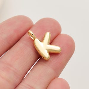 Peut inclure: Un pendentif doré en forme de lettre "K". Le pendentif a une surface lisse et polie et une petite boucle en haut pour l'attacher à une chaîne ou un bracelet. Le pendentif est tenu dans la main d'une personne.