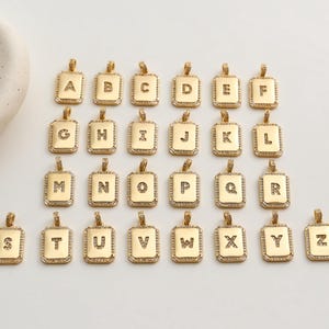 May include: A collection of gold-coloured rectangular pendants, each featuring a different letter of the alphabet from A to Z. The pendants have a square frame with small, clear stones around the edges. The letters are centered.