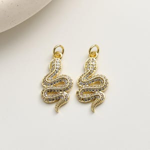 May include: Two gold-coloured snake charms with a loop at the top for hanging. The snake charms are covered in small, clear, sparkling stones. The snake charms are on a white background.