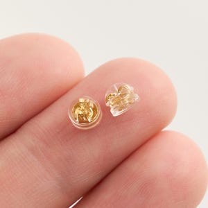 May include: Two gold-colored earring backs, one in a clear plastic casing, are displayed. The earring backs have a butterfly-style design. The image is a close-up shot, highlighting the details of the jewelry findings.