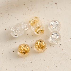 May include: An assortment of earring backs in different designs. The image displays clear, gold-coloured, and silver-coloured earring backs. Some are cylindrical with a spiral pattern, others are round with a post insert.