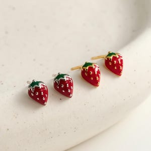 Peut inclure: Quatre boucles d'oreilles en forme de fraise. Deux paires de boucles d'oreilles sont visibles, chacune avec un corps rouge, des mouchetures blanches et un dessus vert. Une paire a des tiges argentées, l'autre dorées.