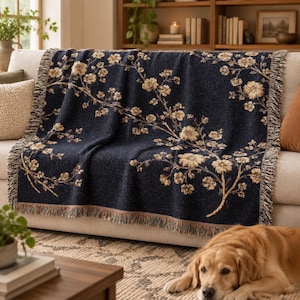 May include: A dark navy blue woven throw blanket featuring a repeating pattern of light beige and tan floral branches. The blanket has a textured finish with fringed edges along the sides, displaying a detailed botanical design across the fabric.