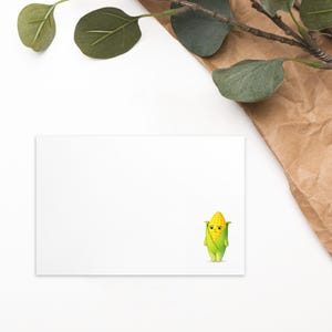 Puede incluir: Una tarjeta rectangular blanca con una ilustración de dibujos animados de un personaje de maíz. El maíz es amarillo con una cáscara verde y tiene una expresión triste. Un papel marrón y hojas verdes están en el fondo.