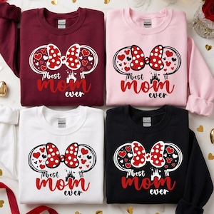 May include: Four sweatshirts in burgundy, pink, white, and black. Each has a design with a red polka-dot bow, hearts, and the text "Best Mom Ever" in red. The design is framed by Mickey Mouse ears.