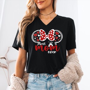 May include: Black V-neck t-shirt with a graphic design featuring a red polka-dot bow, hearts, and the text "Best Mom Ever" in red. The design includes a castle silhouette. The shirt is a casual wear item.