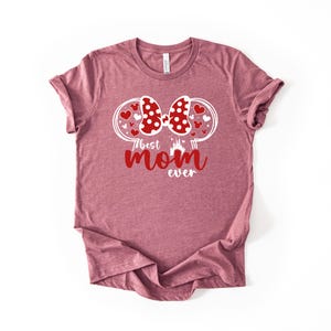 May include: A mauve t-shirt with a graphic design. The design features a red polka-dot bow, heart accents, and the text "best mom ever" in red. A castle silhouette is also included.