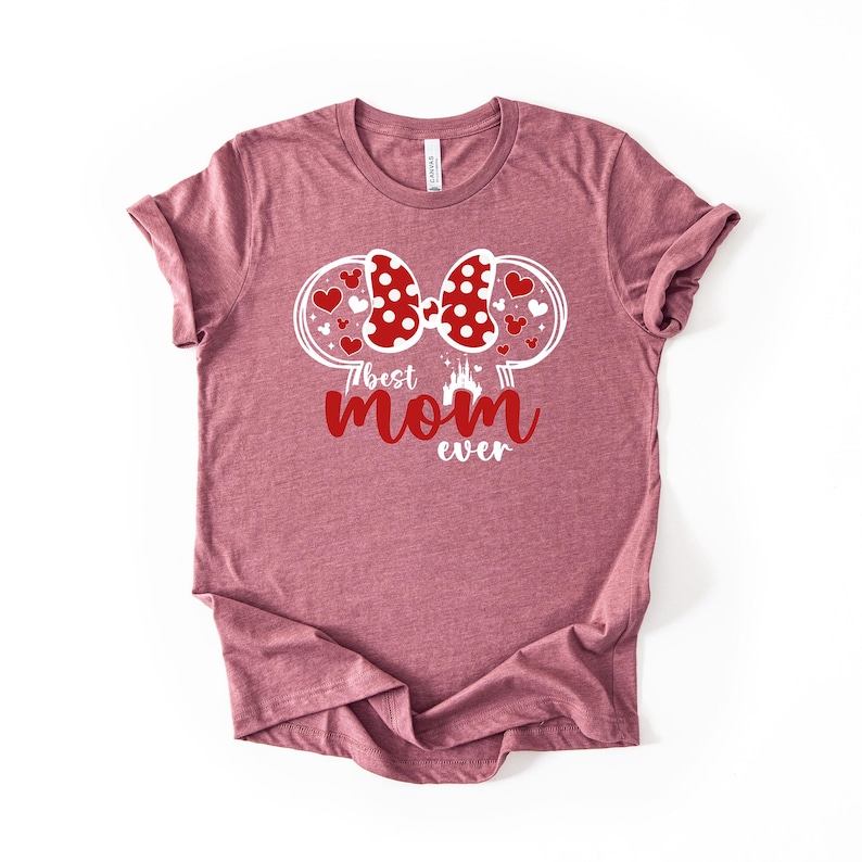 May include: A mauve t-shirt with a graphic design. The design features a red polka-dot bow, heart accents, and the text "best mom ever" in red. A castle silhouette is also included.