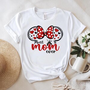 May include: White t-shirt with a red and white polka dot bow design, heart accents, and the text "Best Mom Ever" in red and black. The design includes a castle silhouette. A casual, everyday top.