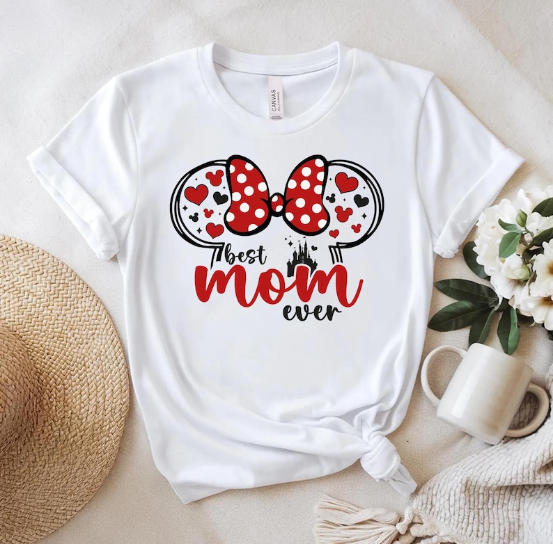 May include: White t-shirt with a red and white polka dot bow design, heart accents, and the text "Best Mom Ever" in red and black. The design includes a castle silhouette. A casual, everyday top.