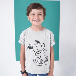 May include: A light gray t-shirt featuring a black and white illustration of Snoopy dancing. The shirt is being worn by a child. The background is a solid teal color.