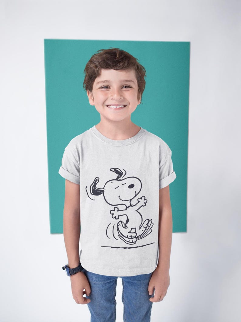 May include: A light gray t-shirt featuring a black and white illustration of Snoopy dancing. The shirt is being worn by a child. The background is a solid teal color.
