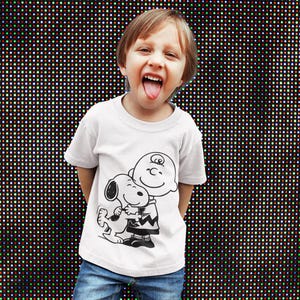 May include: A white t-shirt featuring a black and white graphic of Charlie Brown hugging Snoopy. The child is wearing blue jeans and has their tongue out. The background is a wall of colorful lights.