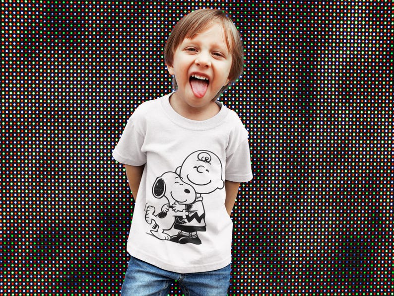 May include: A white t-shirt featuring a black and white graphic of Charlie Brown hugging Snoopy. The child is wearing blue jeans and has their tongue out. The background is a wall of colorful lights.
