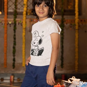 May include: A young person wearing a light gray t-shirt with a black and white cartoon graphic of Snoopy and Woodstock. The person is also wearing blue pants. The background has decorative lights and floral garlands.