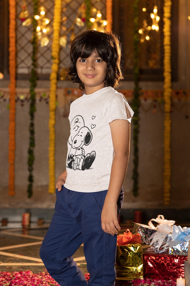 May include: A young person wearing a light gray t-shirt with a black and white cartoon graphic of Snoopy and Woodstock. The person is also wearing blue pants. The background has decorative lights and floral garlands.