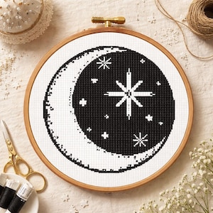 May include: A cross-stitch embroidery hoop featuring a black and white celestial design. The design includes a crescent moon and stars, stitched on white fabric. The wooden hoop is held together with a brass screw. The craft supplies include scissors, thread, and a pin cushion.