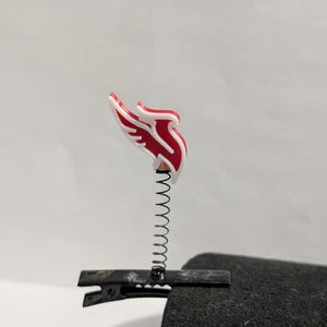 May include: A red and white winged emblem attached to a black metal clip with a silver spring. The emblem is a stylized design, possibly a logo or decorative element. The clip is attached to a dark, textured surface.