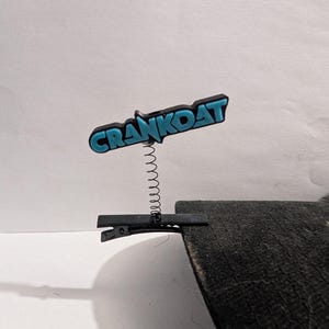 May include: A black hair clip with a spring and a blue and black "CRANKOAT" logo. The clip is attached to a dark grey surface, with a white background.