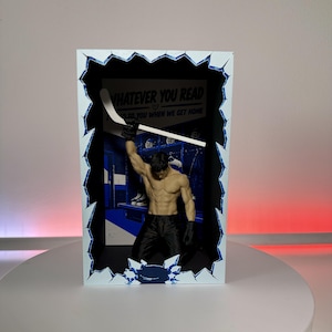 May include: A rectangular shadow box display featuring a shirtless male figure kneeling and holding a white hockey stick aloft. The background depicts a blue locker room scene with the text WHATEVER YOU READ. The frame has a jagged ice-like border.