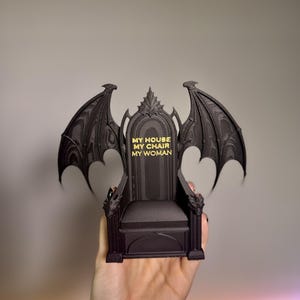 Op de afbeelding: Een zwarte miniatuur troon met drakenvleugel accenten. De troon heeft de tekst "MY HOUSE MY CHAIR MY WOMAN" in gouden letters. De stoel heeft drakenkop armleuningen en een gedetailleerde rugleuning.