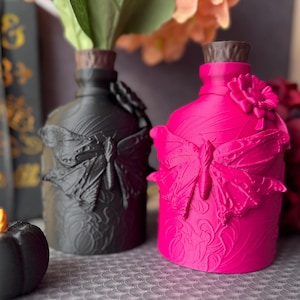 Può includere: Due bottiglie decorative, una nera e una rosa acceso, con decorazioni a farfalla e floreali. Le bottiglie hanno tappi marroni e sono riempite di fiori artificiali. La bottiglia nera ha una finitura opaca, mentre quella rosa ha una finitura lucida.