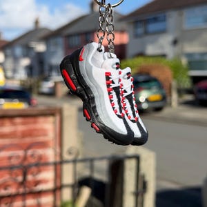 Puede incluir: Un llavero en miniatura con forma de zapatilla deportiva en blanco, negro y gris, con detalles en rojo. El llavero presenta una pequeña réplica de una zapatilla, con cordones y suela texturizada. Está unido a un llavero plateado.