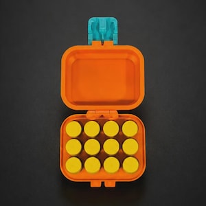 Puede incluir: Un estuche de almacenamiento rectangular de color naranja con tapa abatible y un cierre de color verde azulado. La base interior contiene doce compartimentos circulares pequeños de color amarillo dispuestos en una cuadrícula.