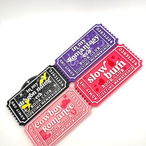 Puede incluir: Cuatro imanes con forma de ticket con los textos "In my Romantasy era", "Slow Burn", "Cowboy Romance" e "In my dragon riding era". Cada imán tiene un color y diseño únicos, con las palabras "Book Club" y "Lifetime Membership".
