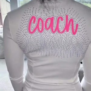 May include: White long-sleeved athletic top with a mock neck and a pink "coach" script across the back. The back of the top is embellished with small, silver-colored rhinestones. The top is a sporty, casual style.