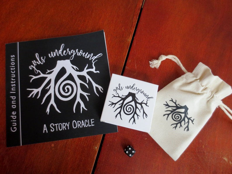 May include: A black and white booklet with the text "girls underground A STORY ORACLE" and a white illustration of a tree root system with a spiral. A white card with the same illustration and text is next to the booklet. A white drawstring bag with the same illustration and text is on a wooden surface. A black die is on the surface.