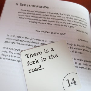 May include: A white card with black text that reads "There is a fork in the road." The card is numbered 14 in the bottom right corner. The card is partially covering a book page with text about a story and a reading.