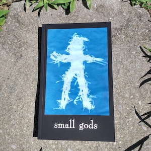 May include: A book titled "small gods" with a blue cover featuring a white silhouette of a person. The person is made of a textured material, possibly fabric or string.