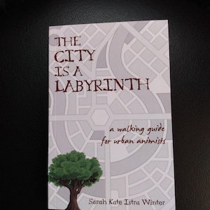 May include: Book cover with a white background and a gray maze pattern. The title "THE CITY IS A LABYRINTH" is in brown text. The subtitle "a walking guide for urban animists" is also in brown text. A green tree is in the lower left corner. The author's name "Sarah Kate Istra Winter" is in brown text at the bottom.