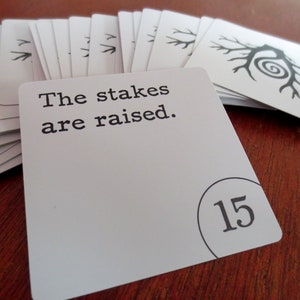 May include: A white card with black text that reads "The stakes are raised."  The card has a black circle with the number 15 in it.  The card is part of a deck of cards.