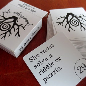May include: A white box with the text "Explore the Power of Story" and "Girls Underground A Story Oracle" with a black and white illustration of a tree with a spiral inside. A white card with the text "She must solve a riddle or puzzle." and the number 29 in a circle.