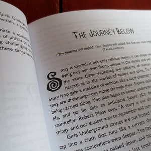 May include: Open book with a white background and black text. The book is titled "The Journey Below" and features a quote: "The journey will unfold. Your destiny will unfold. But first you must begin." (TANGLEWRECK). The text discusses the importance of storytelling and how it can shape reality.