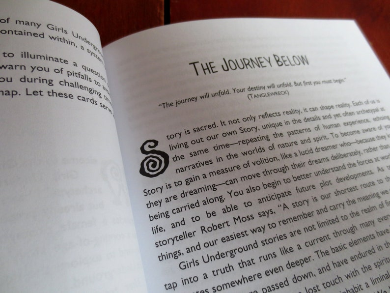 May include: Open book with a white background and black text. The book is titled "The Journey Below" and features a quote: "The journey will unfold. Your destiny will unfold. But first you must begin." (TANGLEWRECK). The text discusses the importance of storytelling and how it can shape reality.
