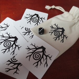 May include: A set of white cards with black tree designs and a black die with white dots. The cards and die are on a wooden surface. The cards are in a white drawstring bag.