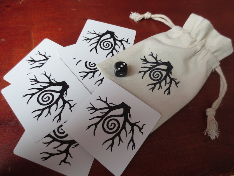 May include: A set of white cards with black tree designs and a black die with white dots. The cards and die are on a wooden surface. The cards are in a white drawstring bag.