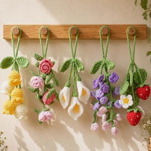 May include: Five hanging crochet ornaments suspended from a wooden rack. The ornaments feature various botanical designs including yellow and white bell-shaped flowers, pink roses, white calla lilies, purple wisteria, and red strawberries with green leaves.