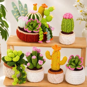 May include: A collection of crocheted cacti and succulents in various shapes and colours, including green, yellow, and pink, potted in small white and dark red crocheted containers. The plants are arranged on a two-tiered wooden display stand.