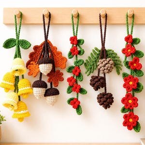 May include: Five hanging crochet ornaments featuring seasonal motifs: yellow bell-shaped flowers with green leaves, brown and cream acorns with orange leaves, a red flower vine, brown pinecones with green fern leaves, and a red flower garland.